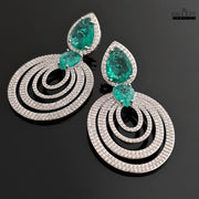 A pair of DHAATU JEWELRY emerald green and pave diamond chandelier earrings, shown at an angle on a black reflective surface.
