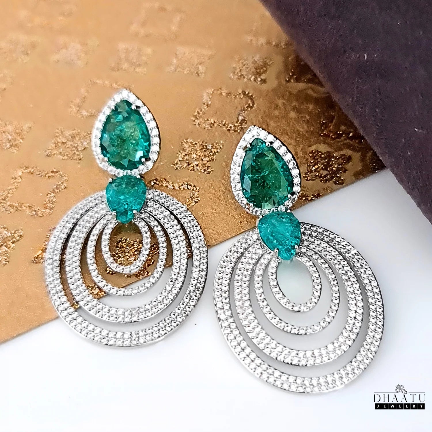 A close-up view of a pair of DHAATU JEWELRY emerald green and pave diamond chandelier earrings, on a textured background.