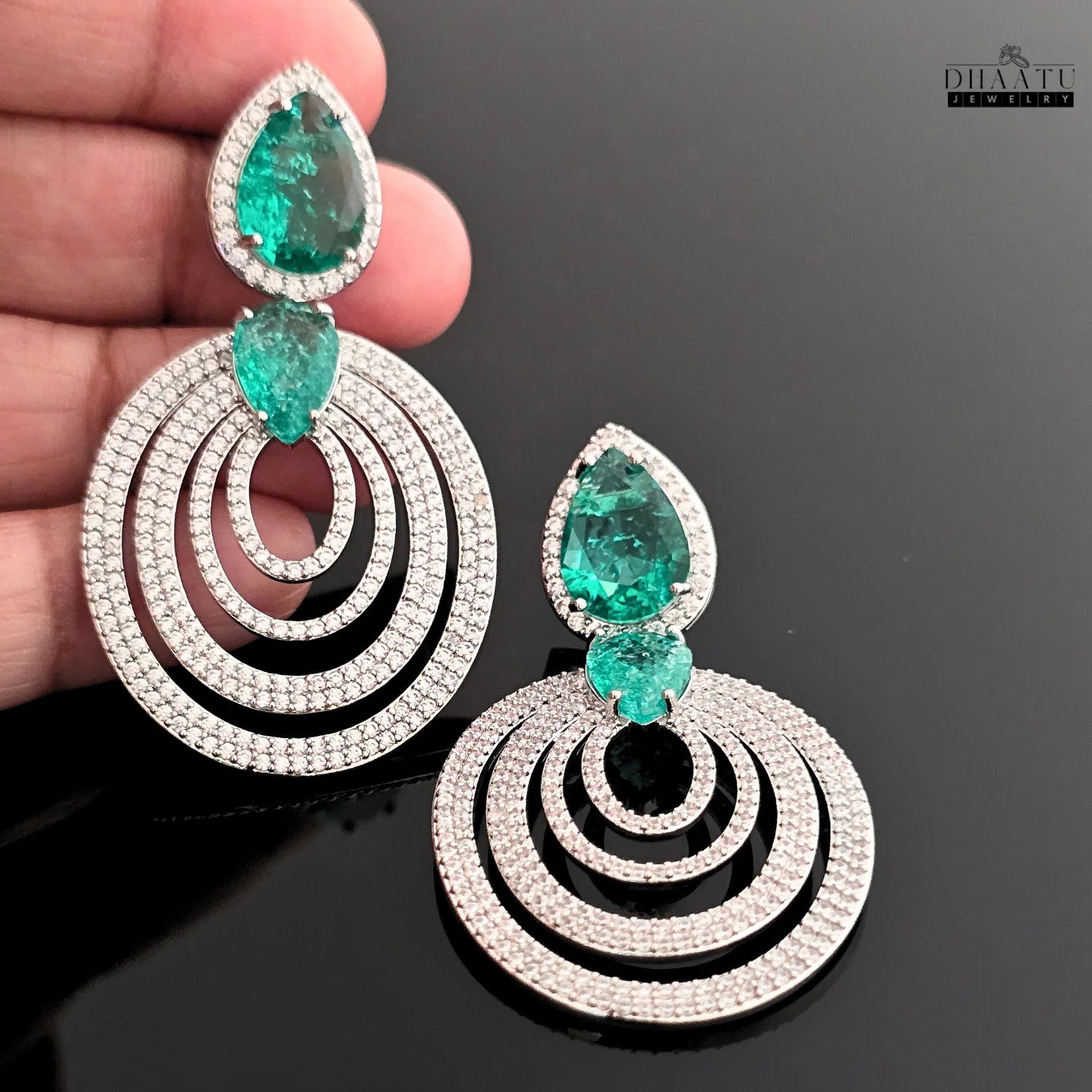 A person holding a pair of DHAATU JEWELRY emerald green and pave diamond chandelier earrings, with a black background.