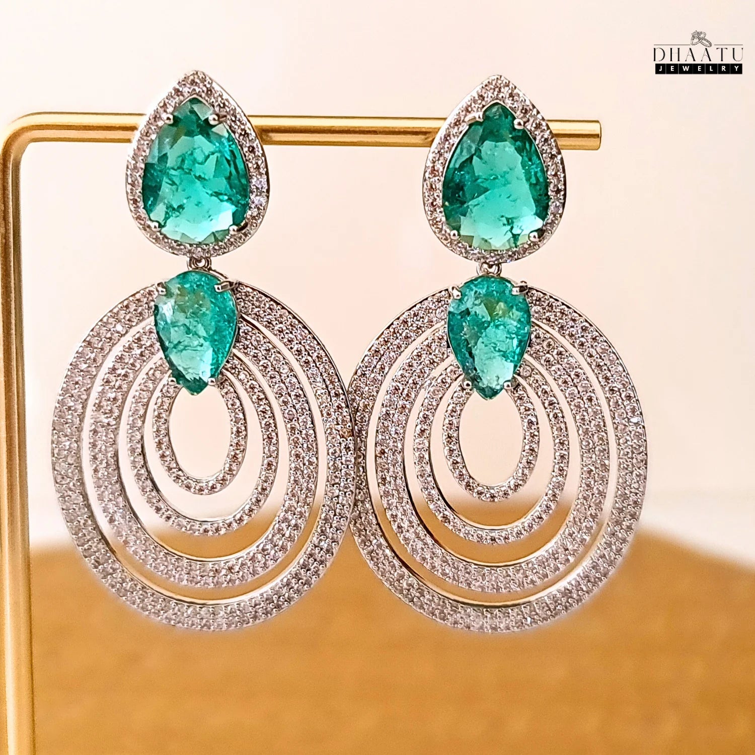 A pair of DHAATU JEWELRY emerald green and pave diamond chandelier earrings displayed on a gold-toned jewelry stand.