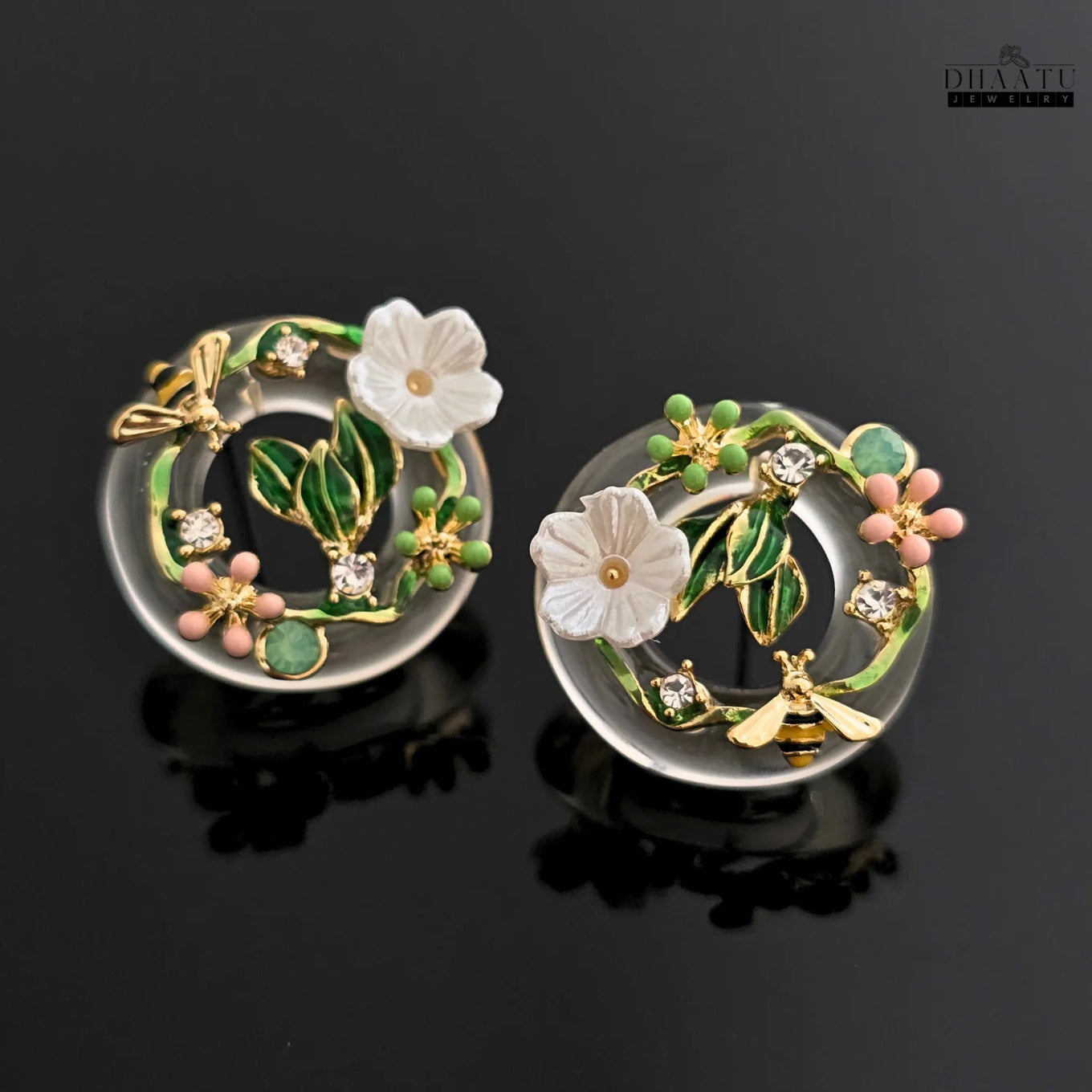 A pair of DHAATU JEWELRY enamel floral bee stud earrings displayed on a reflective black background, emphasizing the clear resin and vibrant colors.