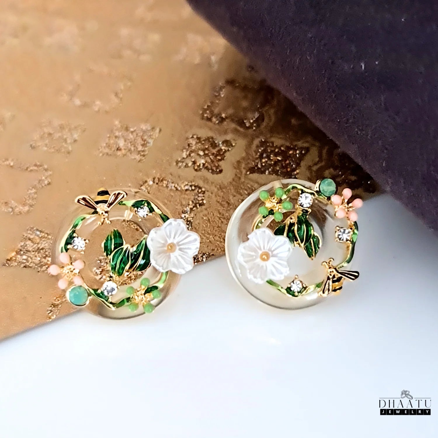 A pair of DHAATU JEWELRY floral and bee stud earrings on a textured gold and brown background, highlighting their unique design.