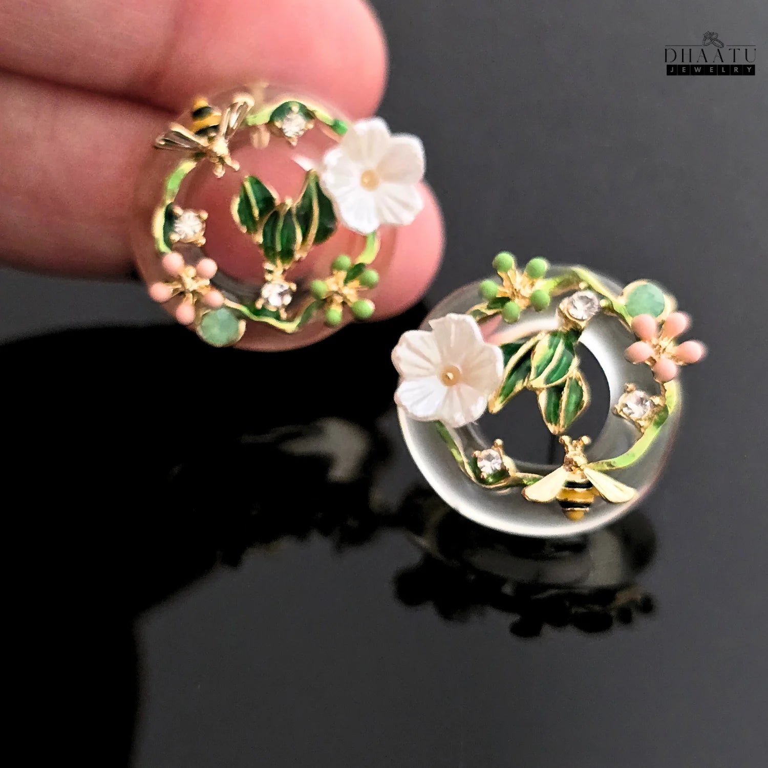 A detailed close-up of a pair of DHAATU JEWELRY floral and bee stud earrings, showcasing the intricate enamel, cubic zirconia, and shell-like flower details.