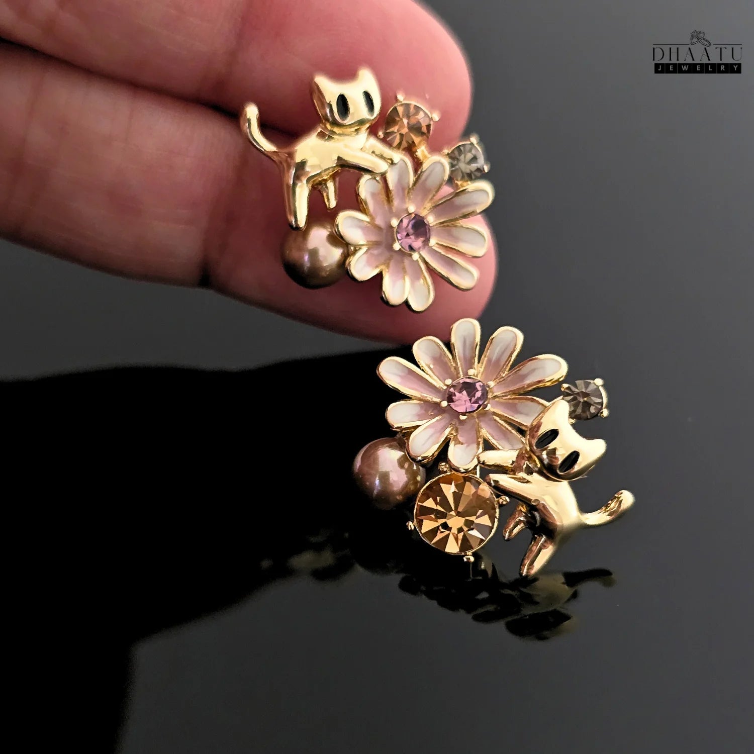 A hand holding the gold cat and floral earrings, providing a clear view of the design and craftsmanship.
