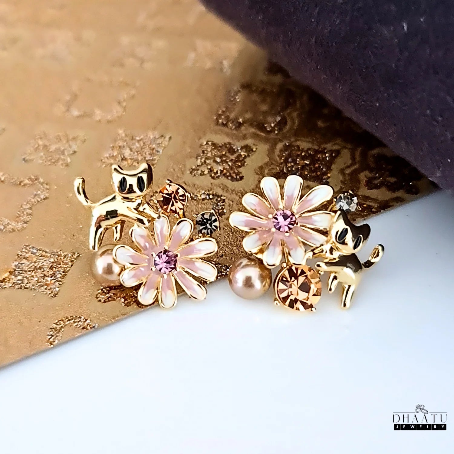 A product shot of the gold cat and floral earrings, featuring a pink enamel flower and a mix of amber and pink crystals.