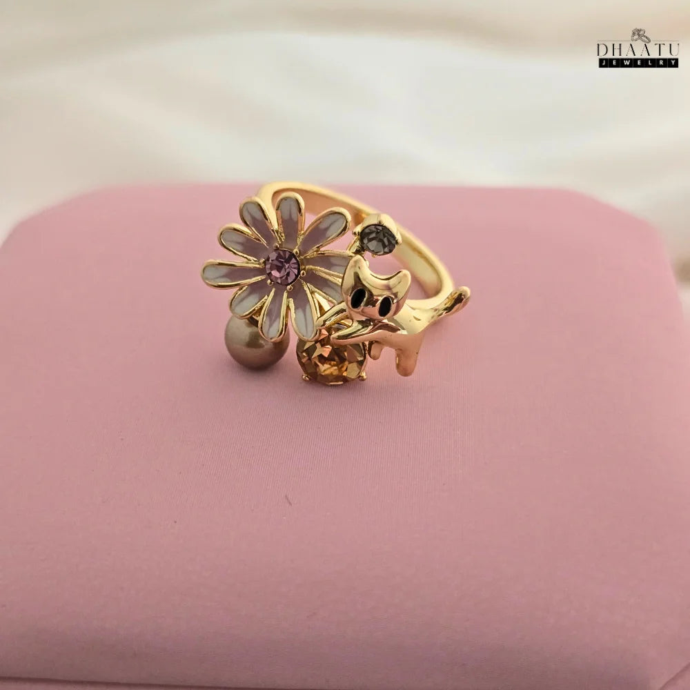 The adjustable gold-toned ring with a playful cat and flower design, displayed on a pink surface to showcase the craftsmanship and warm tones.