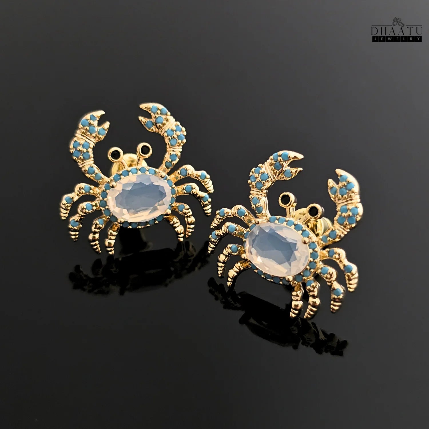 Pair of gold-toned crab earrings with turquoise and white zircon stones on a glossy black background