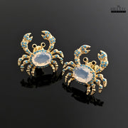 Pair of gold-toned crab earrings with turquoise and white zircon stones on a glossy black background
