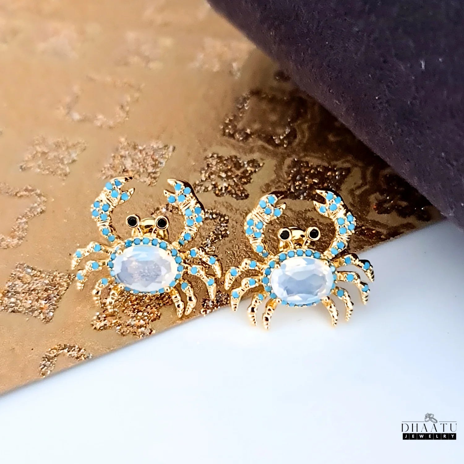 Pair of gold-toned crab earrings with turquoise and white zircon stones on a textured gold and white background.