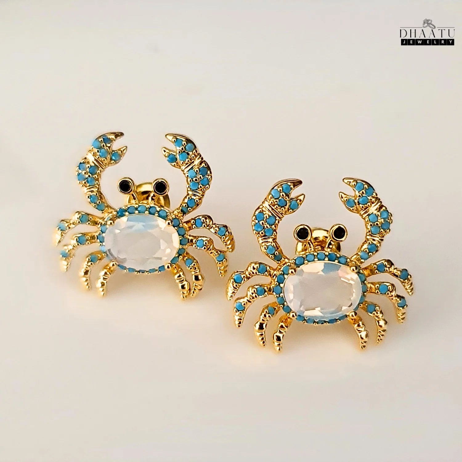 Pair of gold-toned crab earrings with turquoise and white zircon stones on a white background.