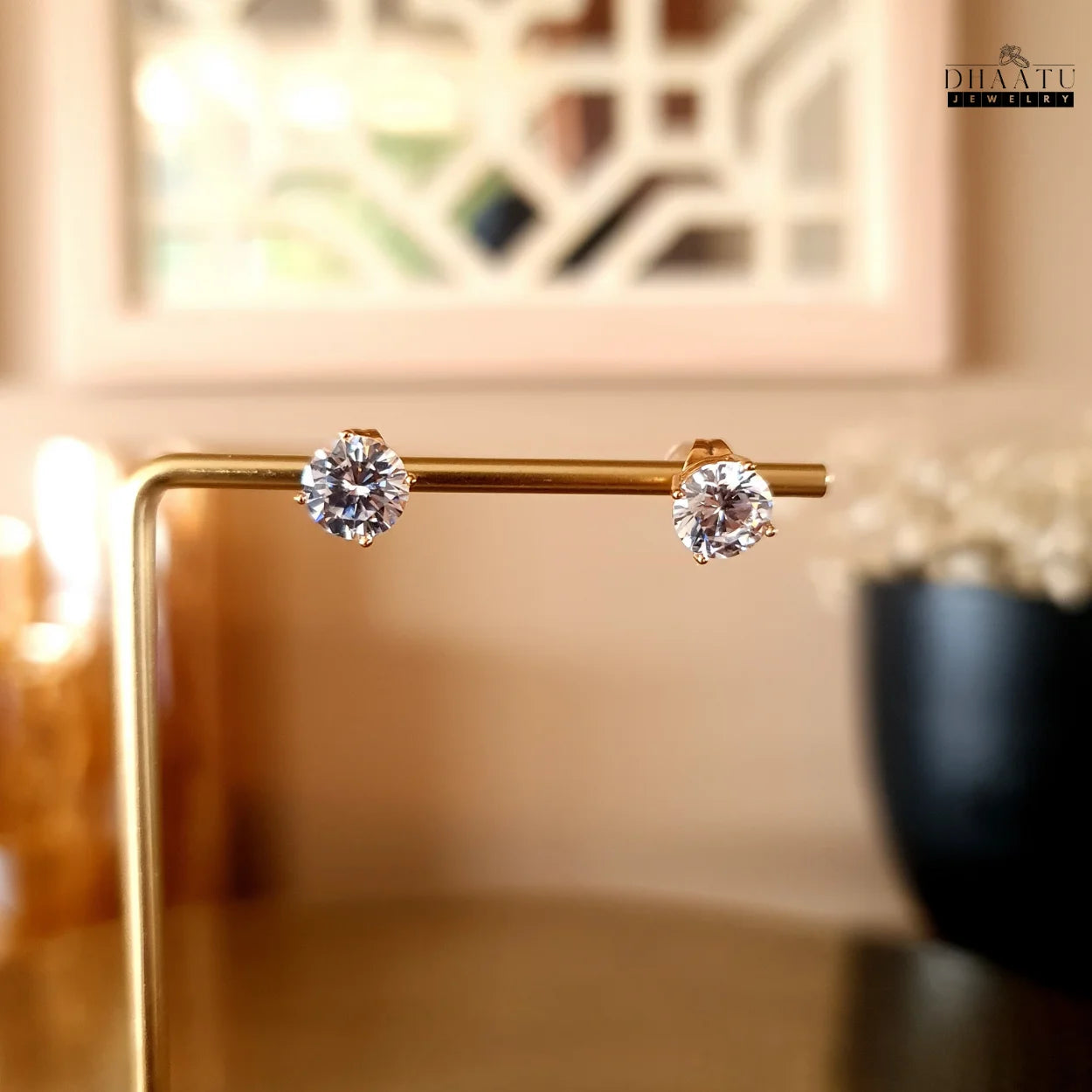 Dhaatu Jewelry gold-tone solitaire stud earrings with clear cubic zirconia gems, elegantly displayed on a golden stand against a soft, blurred background.