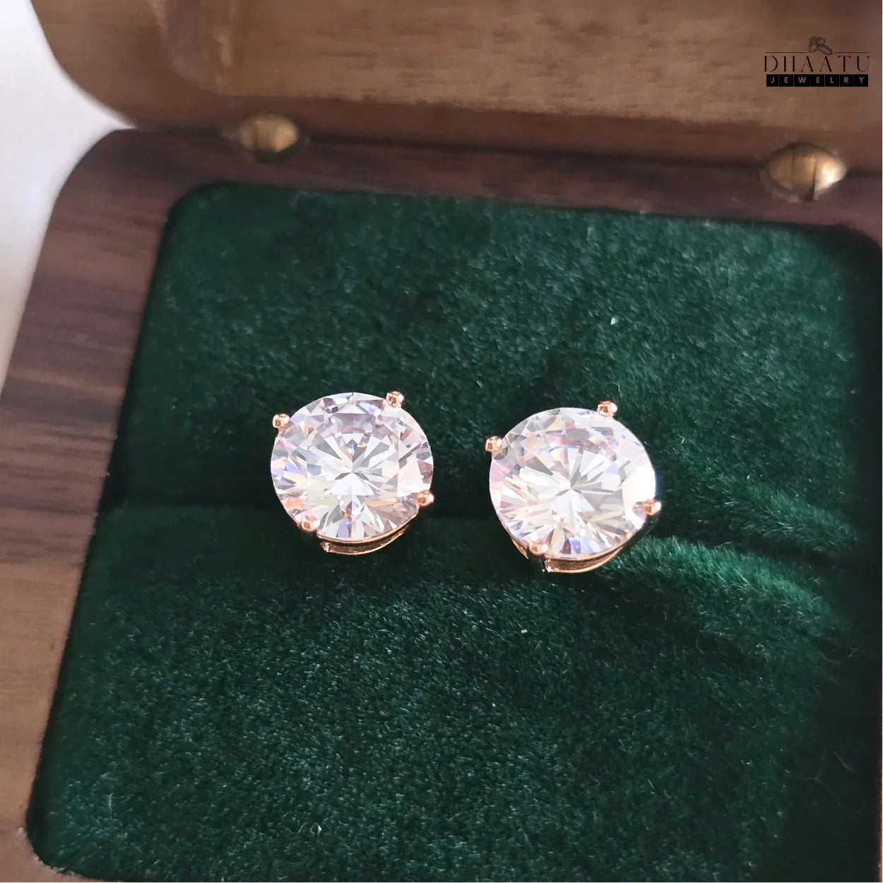 Dhaatu Jewelry gold-tone solitaire stud earrings with shimmering cubic zirconia, displayed open in a dark green velvet box, emphasizing their elegant presentation.

