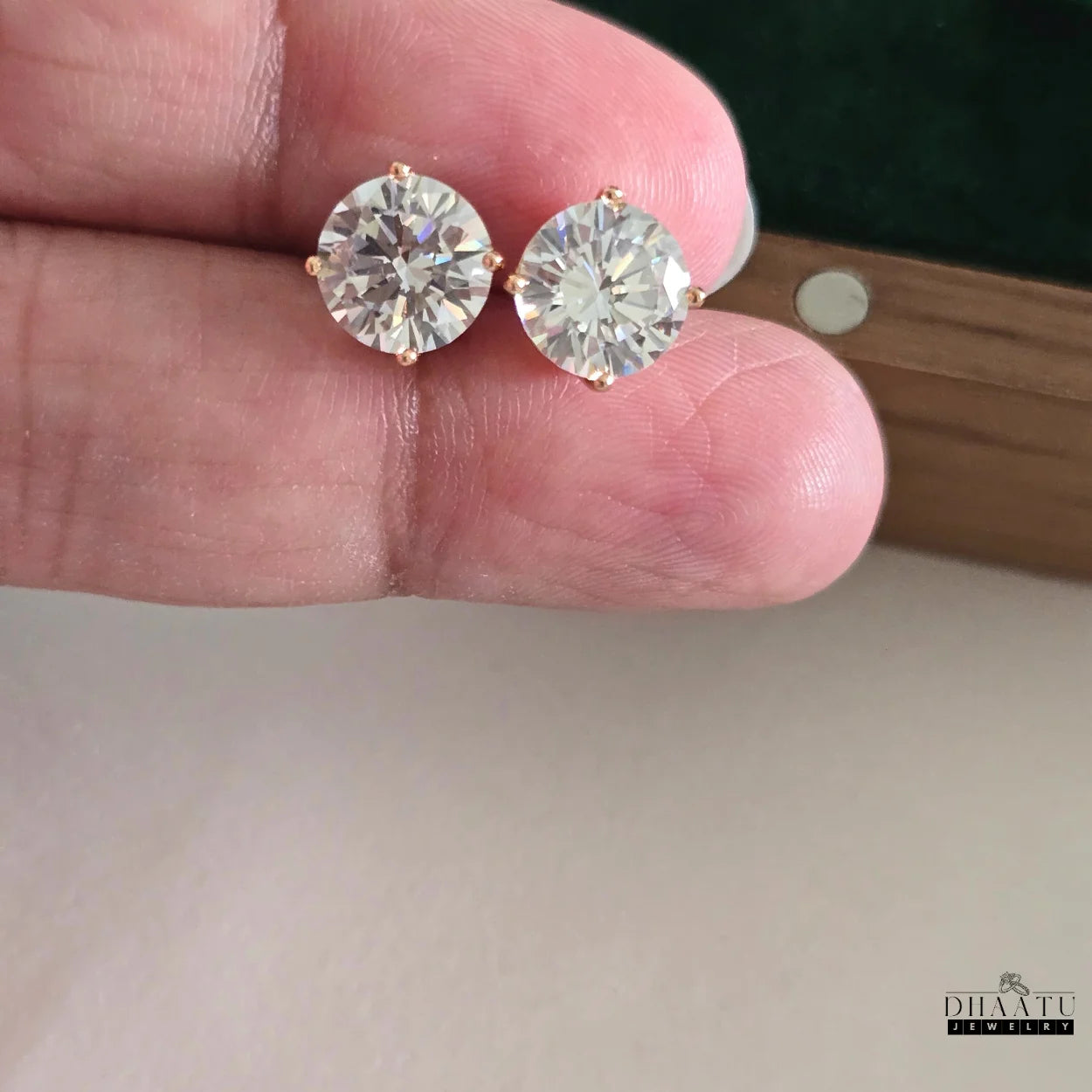 A pair of Dhaatu Jewelry gold-tone solitaire stud earrings with brilliant cubic zirconia, held delicately in a hand against a soft, light background, highlighting their appeal.