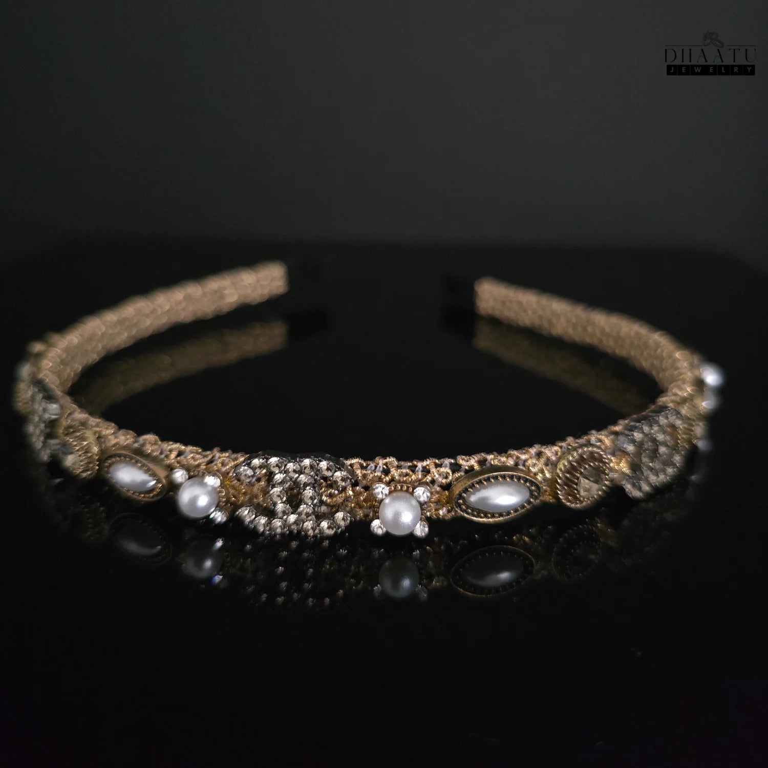 Close-up of Dhaatu Jewelry golden woven hairband with pearls and crystals on a dark reflective surface, showcasing intricate western design.