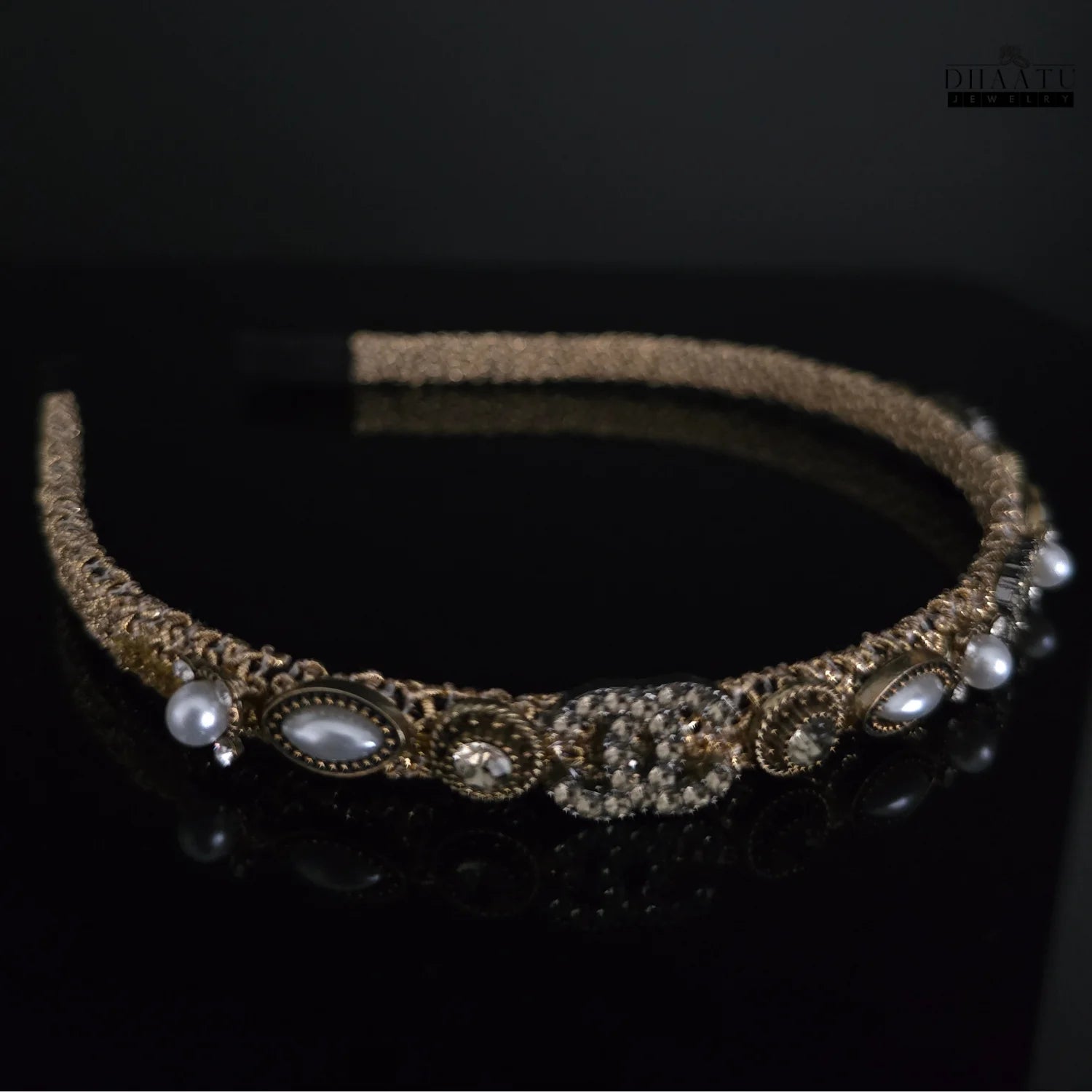 Side angle view of Dhaatu Jewelry modern golden woven hairband with pearl and crystal details on a dark reflective surface.