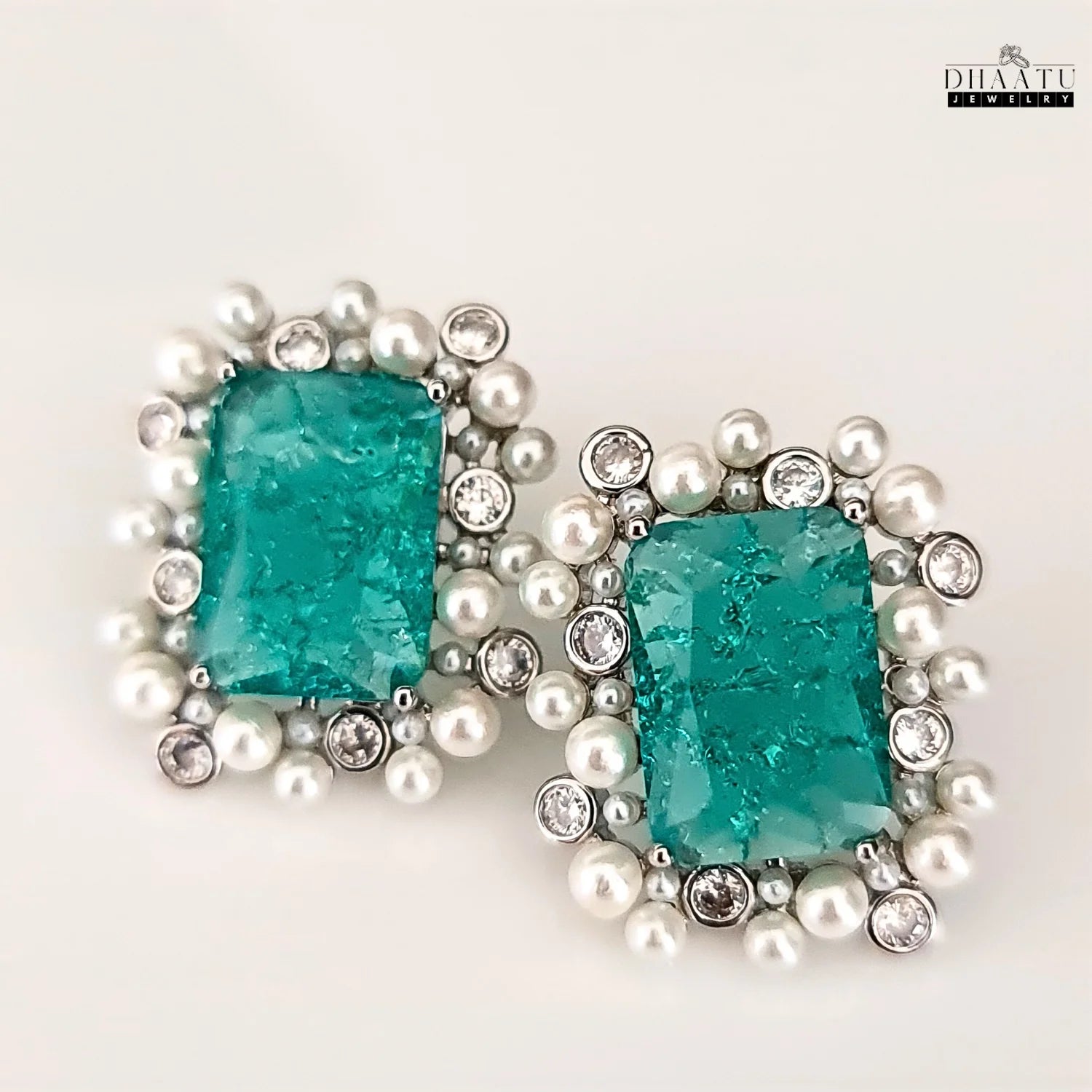 Pair of earrings with turquoise stones and pearl accents on a white background, branded 'Dhaatu'.