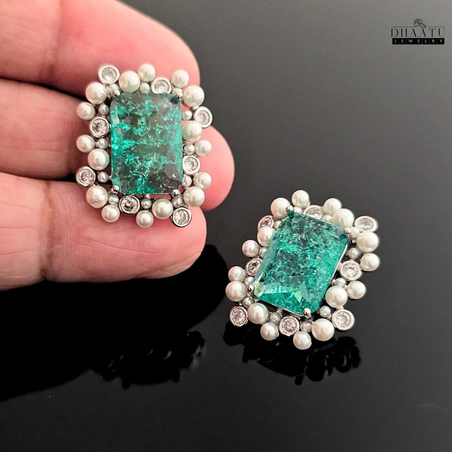 Pair of earrings with green gemstones and pearl-like accents held by a hand on a dark background.