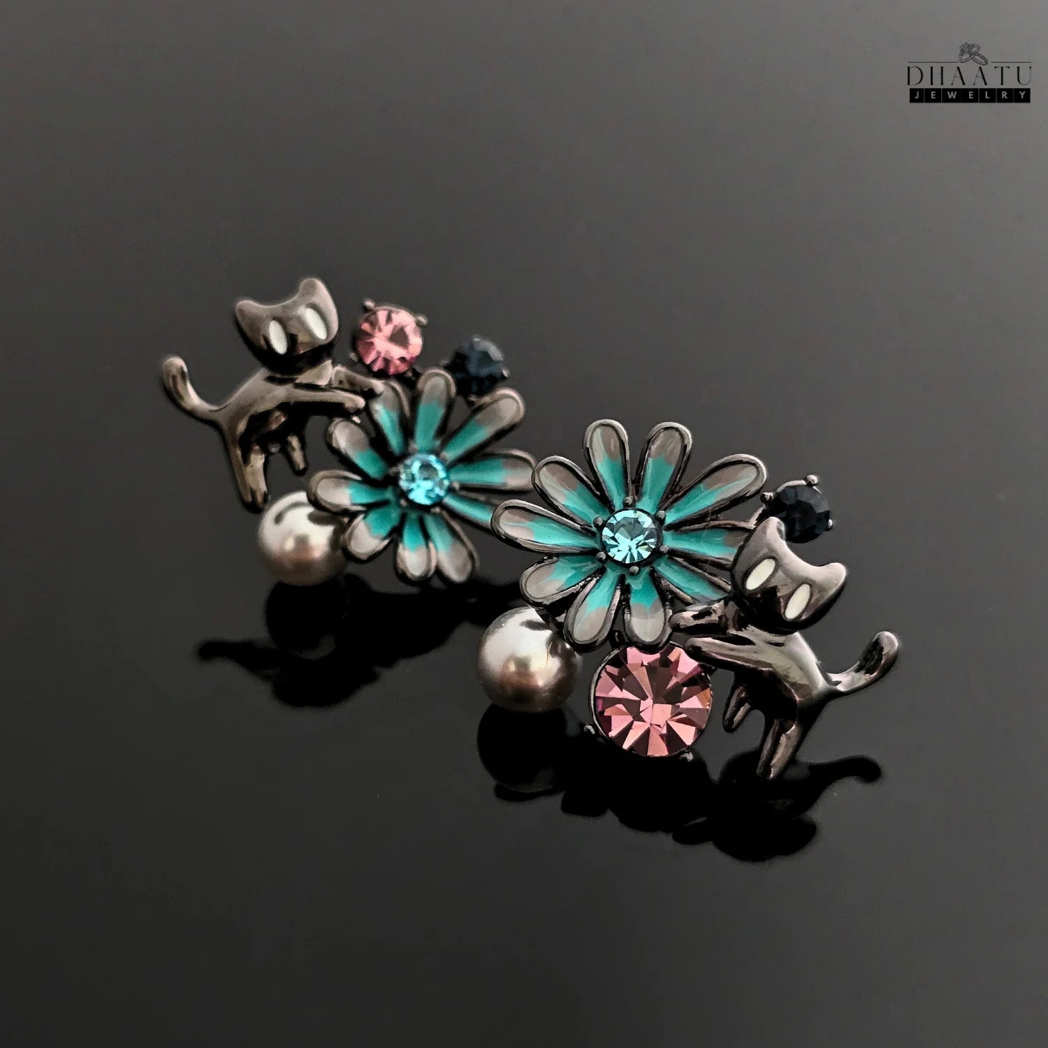 The gunmetal cat and floral earrings placed on a reflective black surface, emphasizing the shine and intricate details.