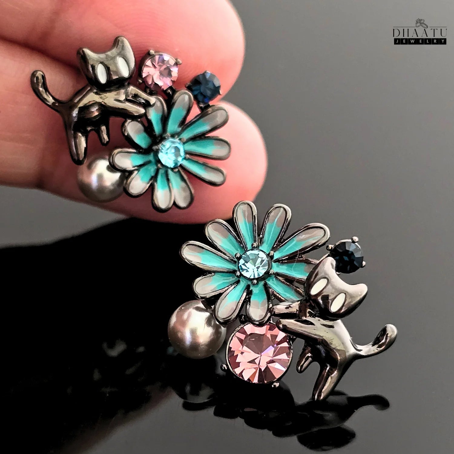 A hand holding up the gunmetal cat and floral earrings, showcasing the detail and scale of the jewelry against a dark background.