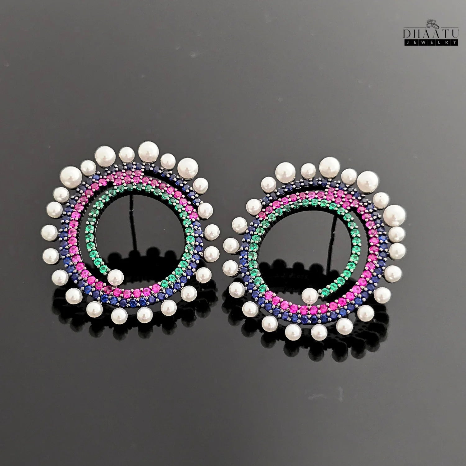 A pair of Dhaatu Jewelry multi-color spiral earrings with blue, pink, and green gemstones and a pearl border, shown on a reflective black surface.