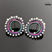 A pair of Dhaatu Jewelry multi-color spiral earrings with blue, pink, and green gemstones and a pearl border, shown on a reflective black surface.