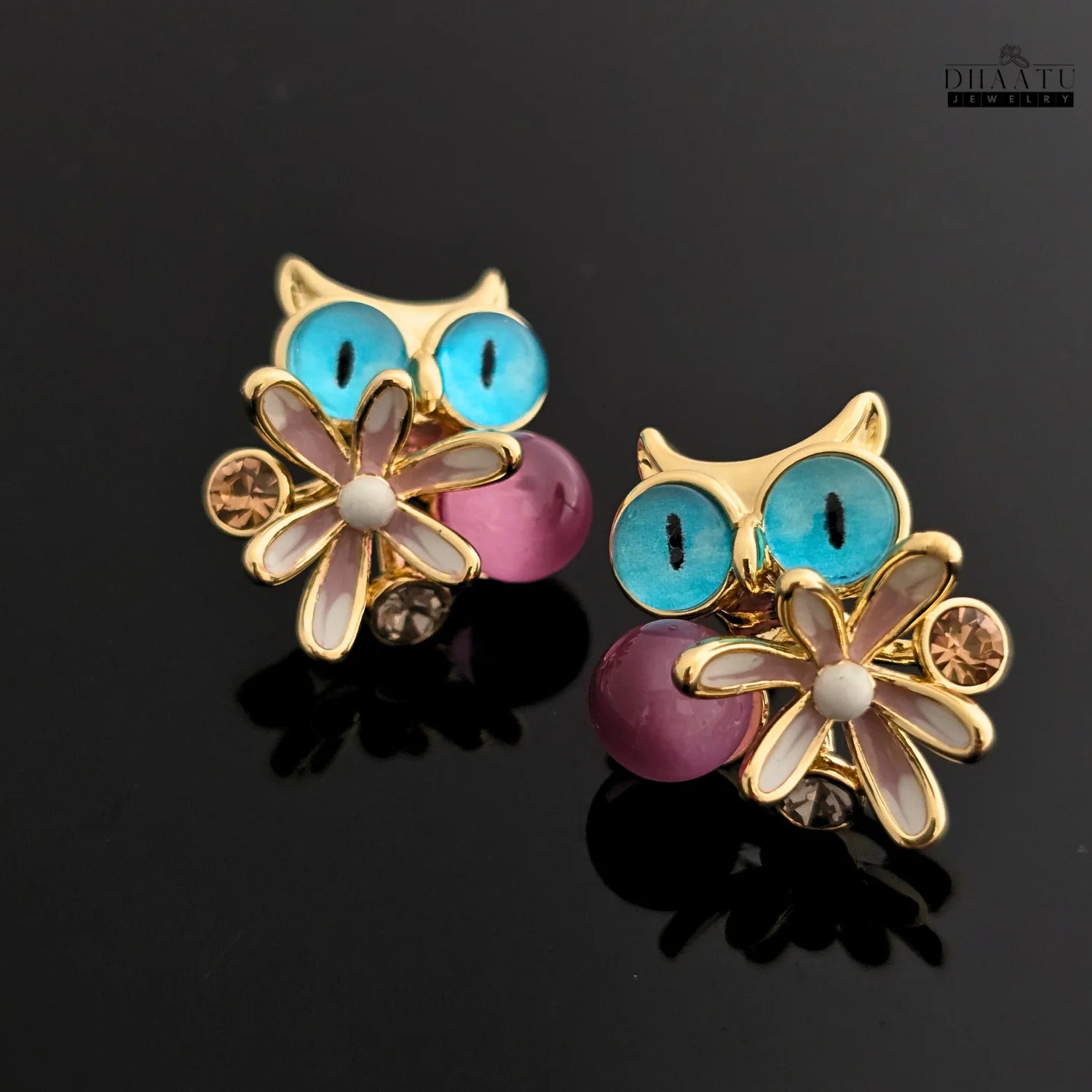 Pair of Dhaatu Jewelry owl stud earrings with blue eyes and pink gemstones on a reflective black background.