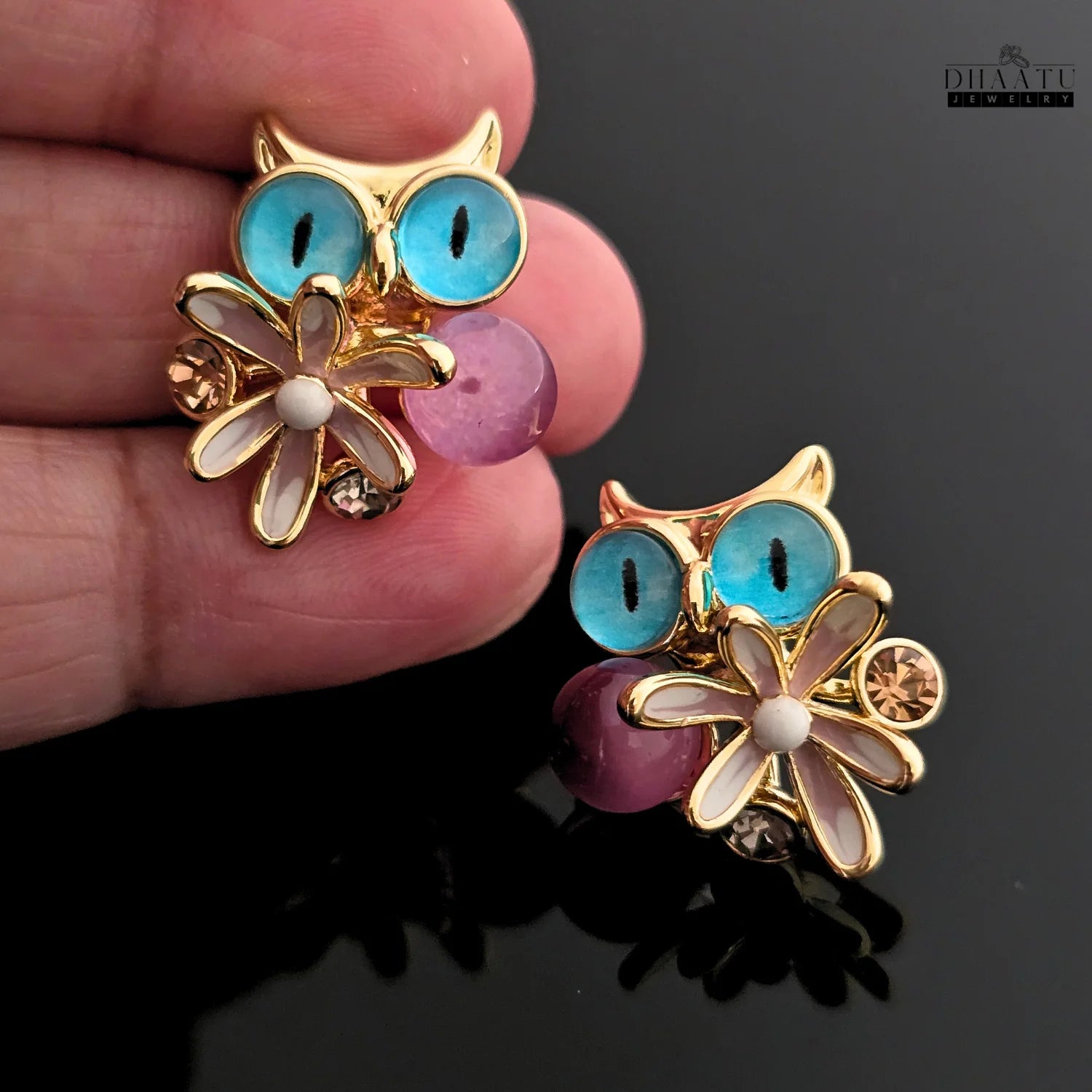 Hand holding one of the Dhaatu Jewelry owl stud earrings with blue eyes, showing the detail up close.