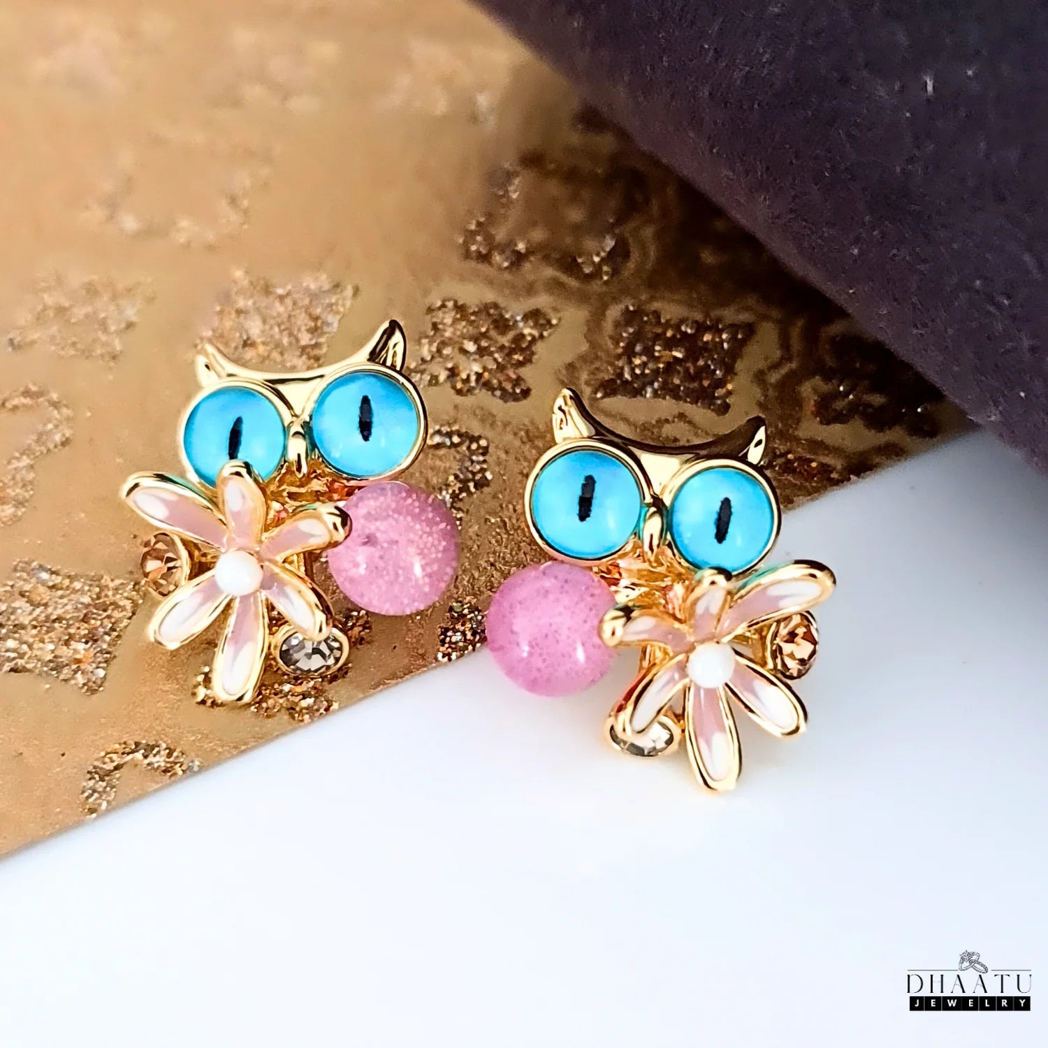 Dhaatu Jewelry gold-toned owl stud earrings with light blue eyes and pink enamel flower on a gold textured background.