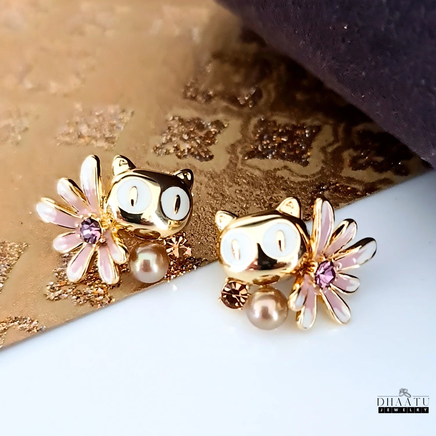 Dhaatu Jewelry gold-toned owl stud earrings with white eyes and mocha pearl on a styled gold and brown background.