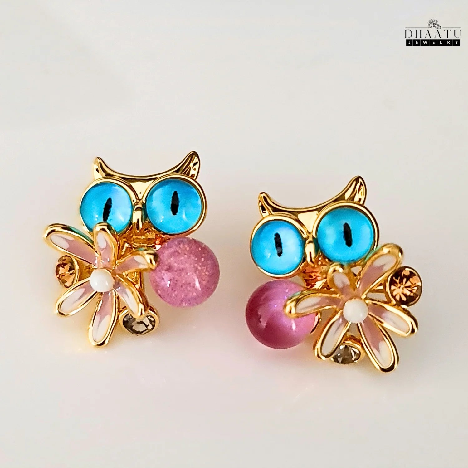 Close-up of Dhaatu Jewelry owl stud earrings with blue eyes, pink flower, and pink gemstone on a white background.
