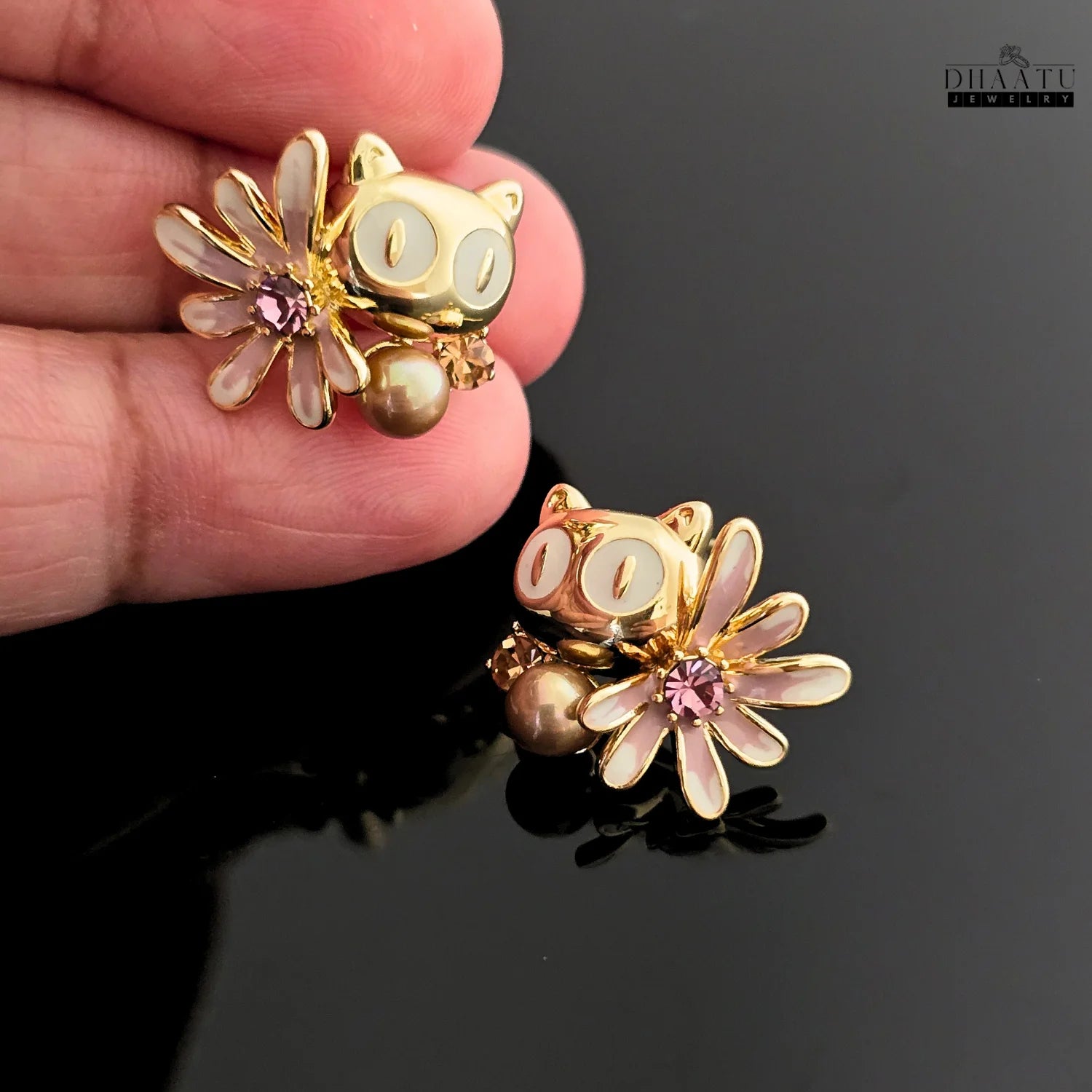 Hand holding one of the Dhaatu Jewelry gold-toned owl stud earrings with white eyes and a pink enamel flower.