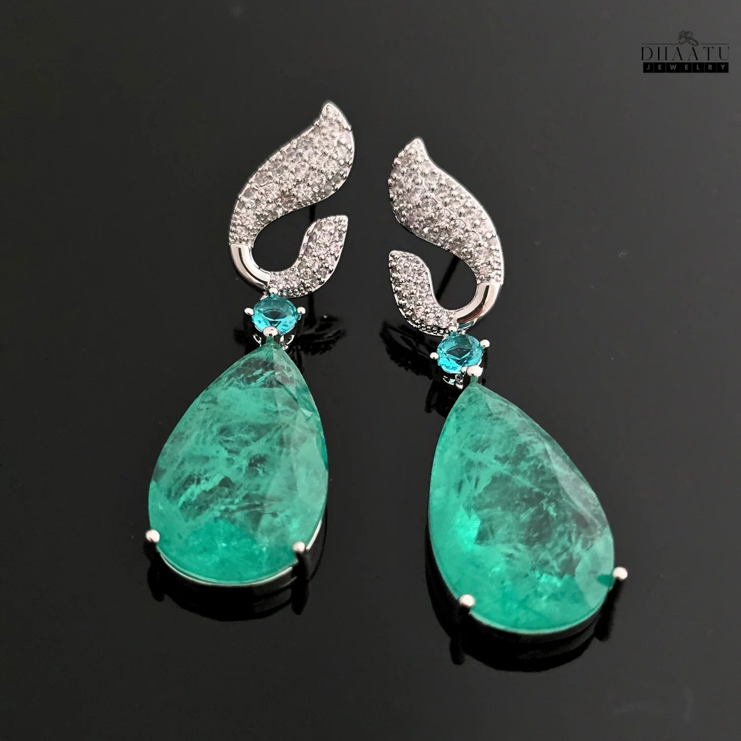 Dhaatu Jewelry green Paraiba stone dangle earrings with white rhodium plating on a reflective black background.

