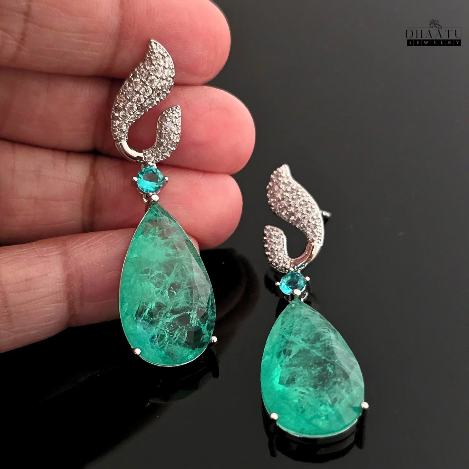 A hand holding up a Dhaatu Jewelry Paraiba green stone dangle earring, showing its size and sparkle.