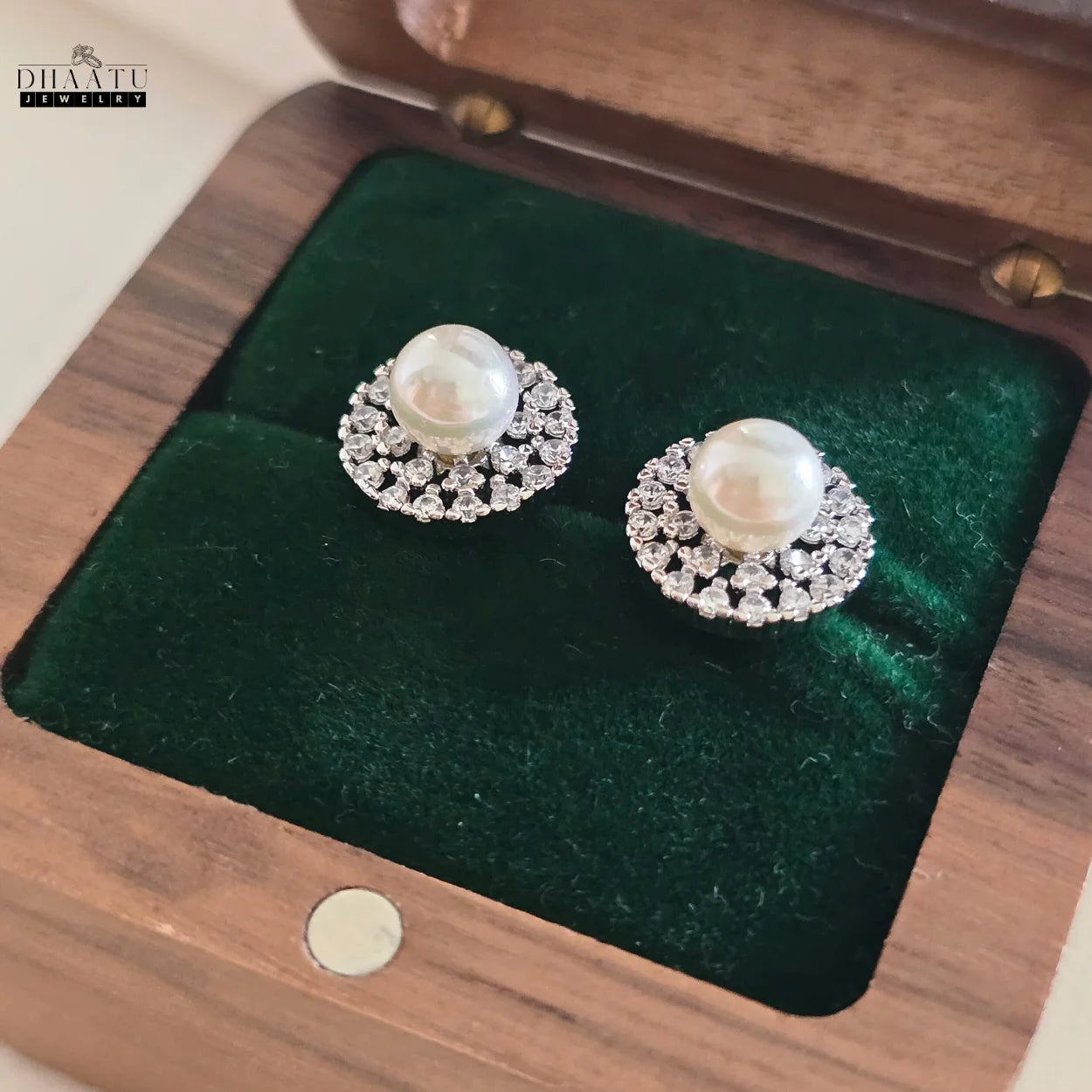Pair of Dhaatu Jewelry pearl and zircon stud earrings placed in a wooden jewelry box with green velvet lining.

