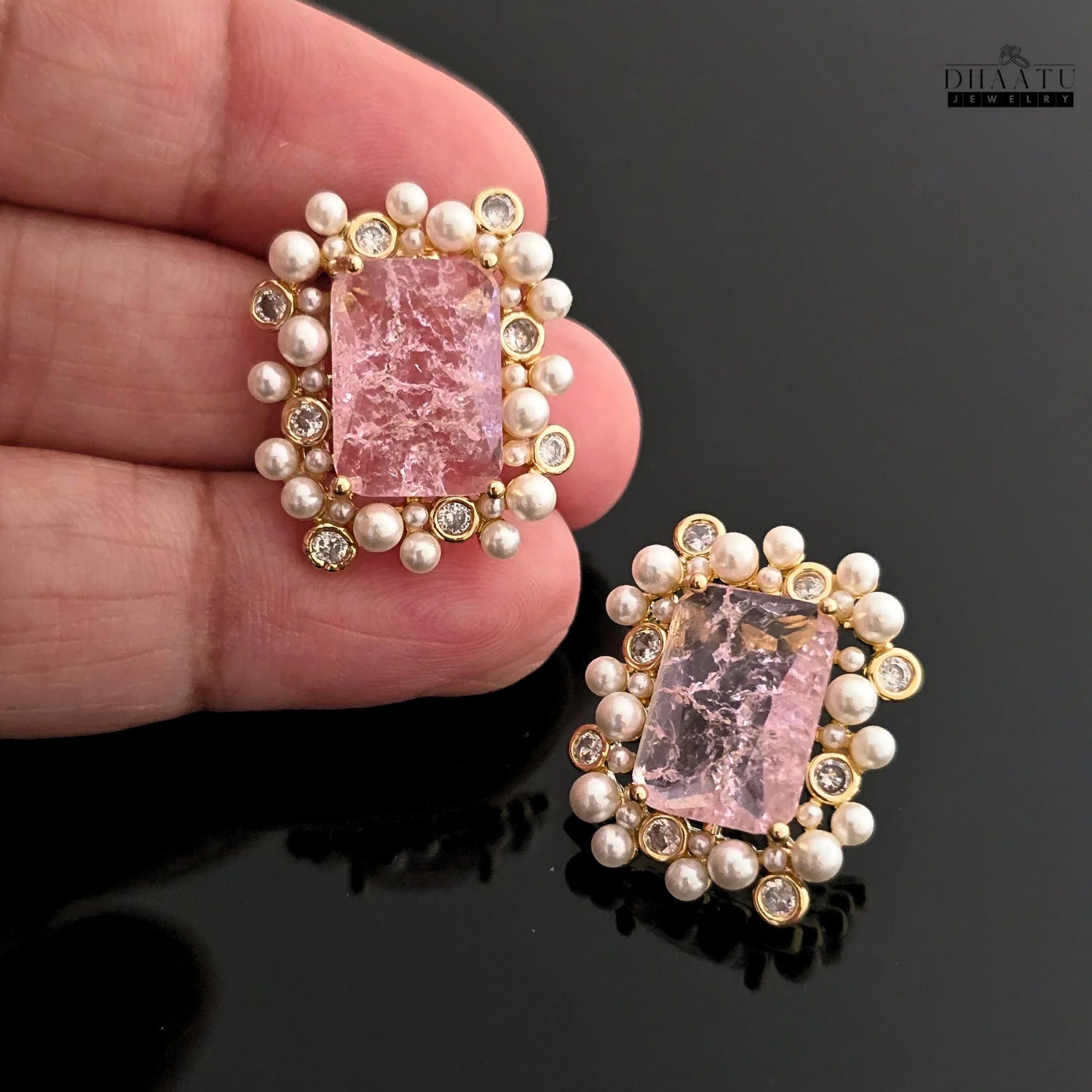 Gold earrings with pink gemstones held by a hand on a dark background, branded 'Dhaatu'.