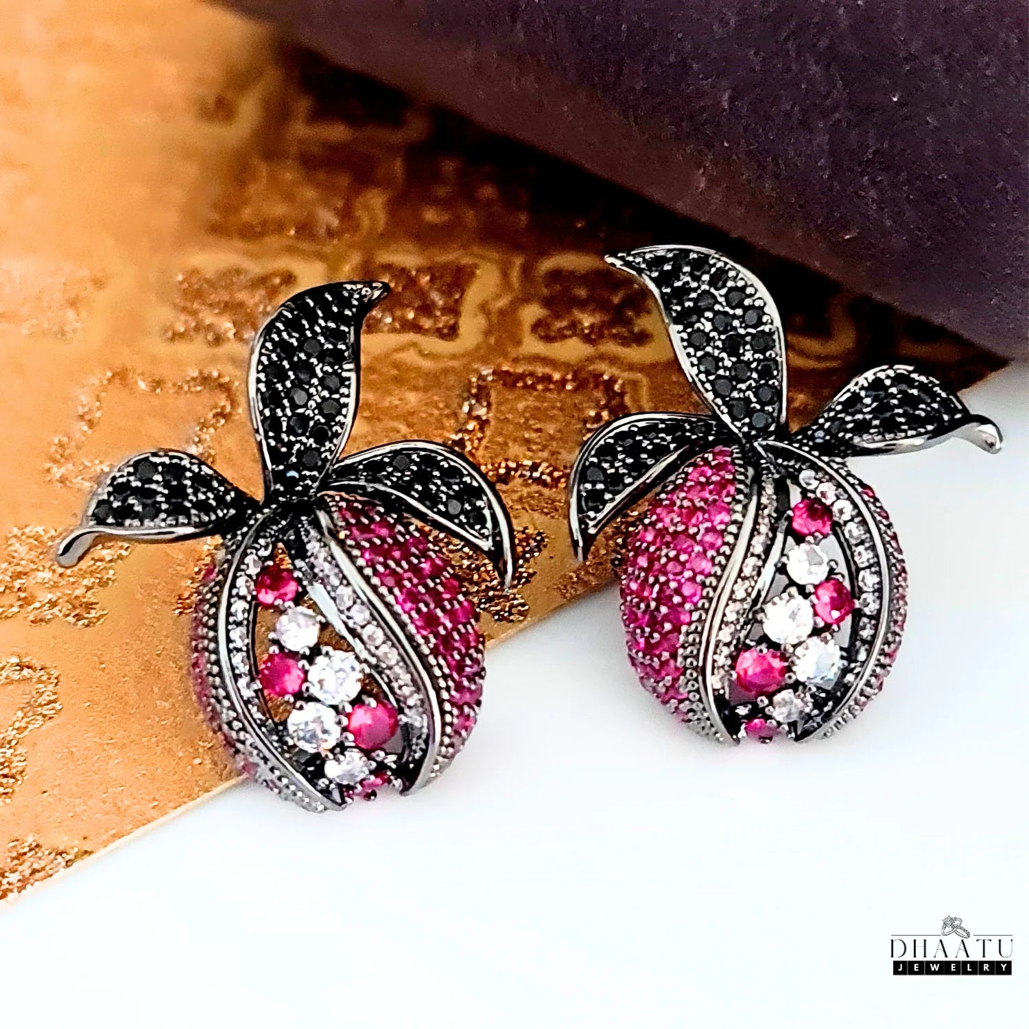 Pomegranate-inspired stud earrings with black and pink zircons, displayed against an artistic gold and purple textured background.