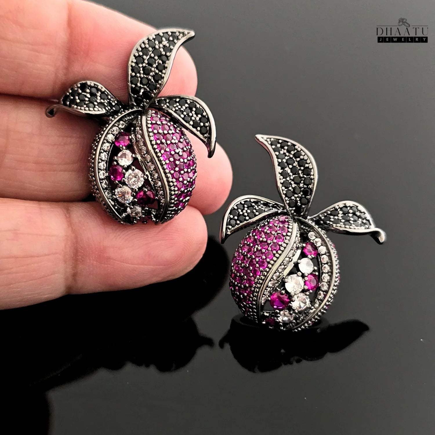 Close-up view of a hand holding a pair of pomegranate stud earrings, showcasing the intricate details of the pink, white, and black zircon stones.
