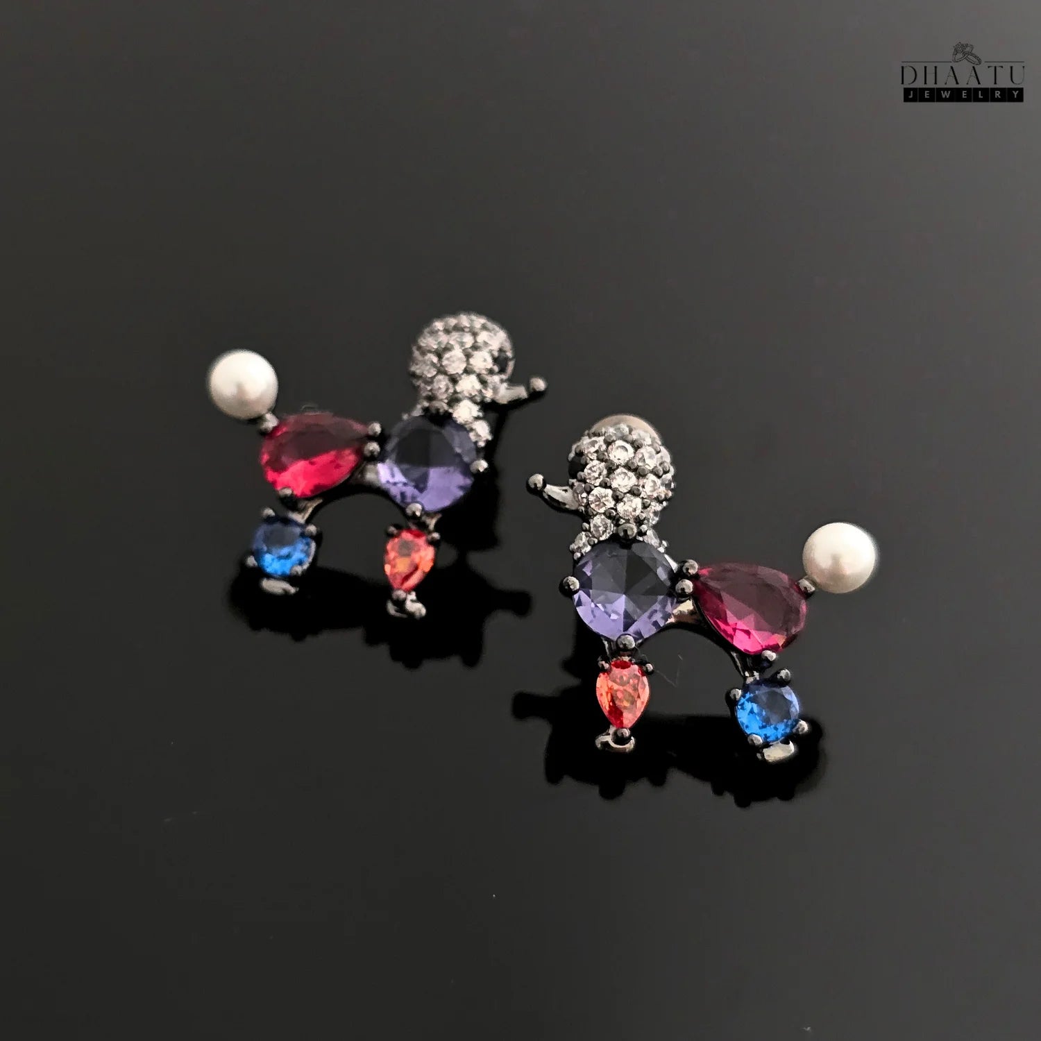 A pair of multicolor poodle-shaped earrings with red, blue, and purple crystals on a reflective black background.