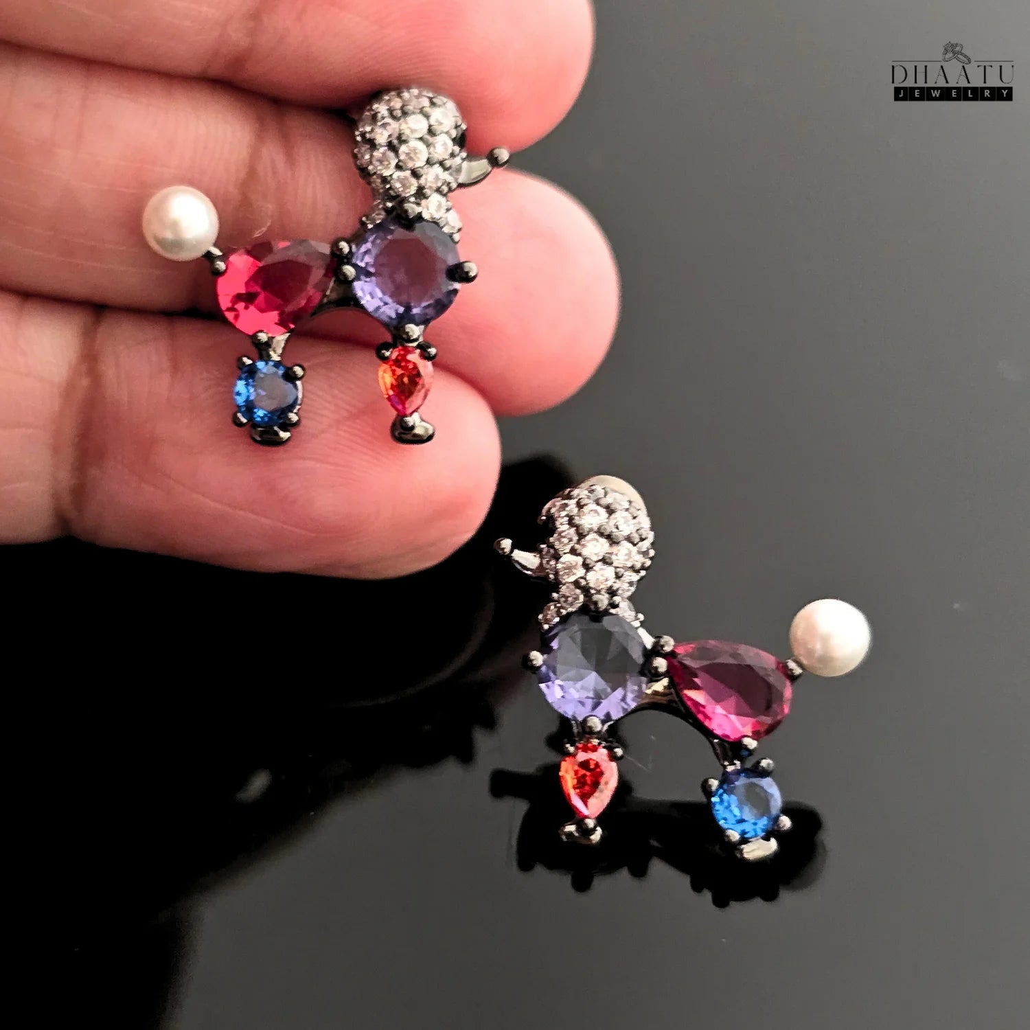 Hand holding a pair of vibrant multicolor poodle earrings with crystals and faux pearls on a black surface.