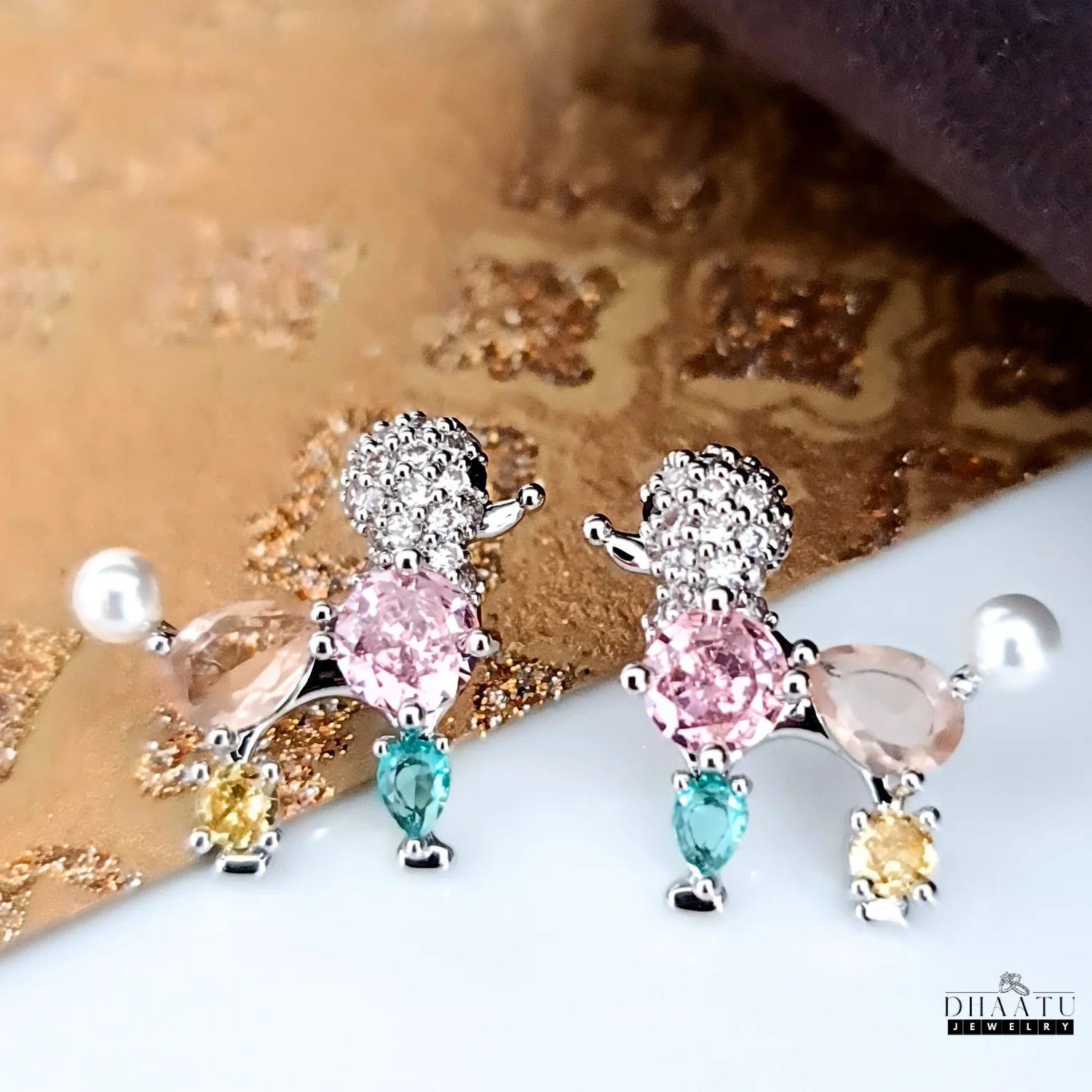 A close-up shot of the pastel poodle earrings with crystals and faux pearls on a textured gold and white surface.