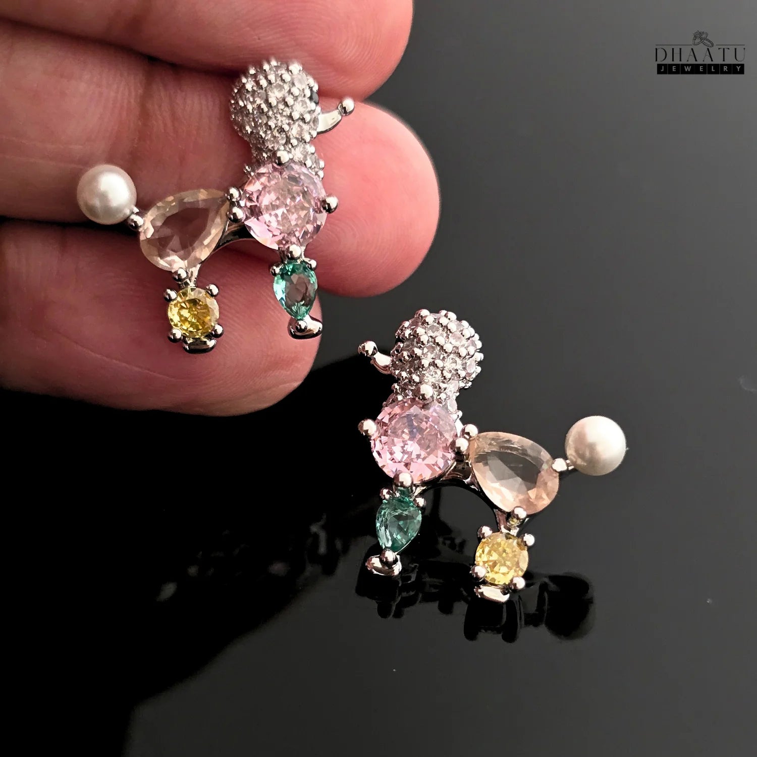 Hand holding a pair of pastel poodle-shaped earrings with crystals and faux pearls on a reflective black surface.