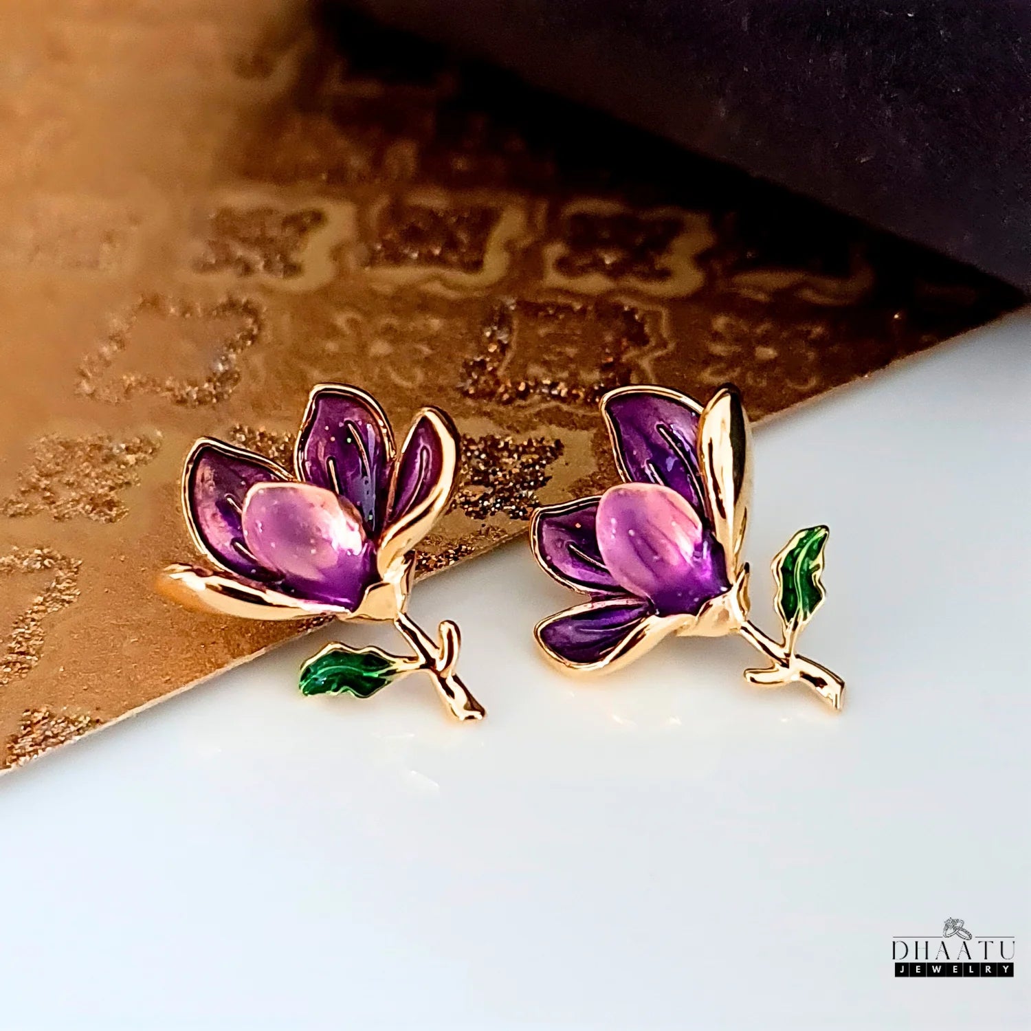 A pair of Dhaatu Jewelry purple floral enamel earrings placed on a textured gold and white background, showcasing their vibrant colors and intricate details.

