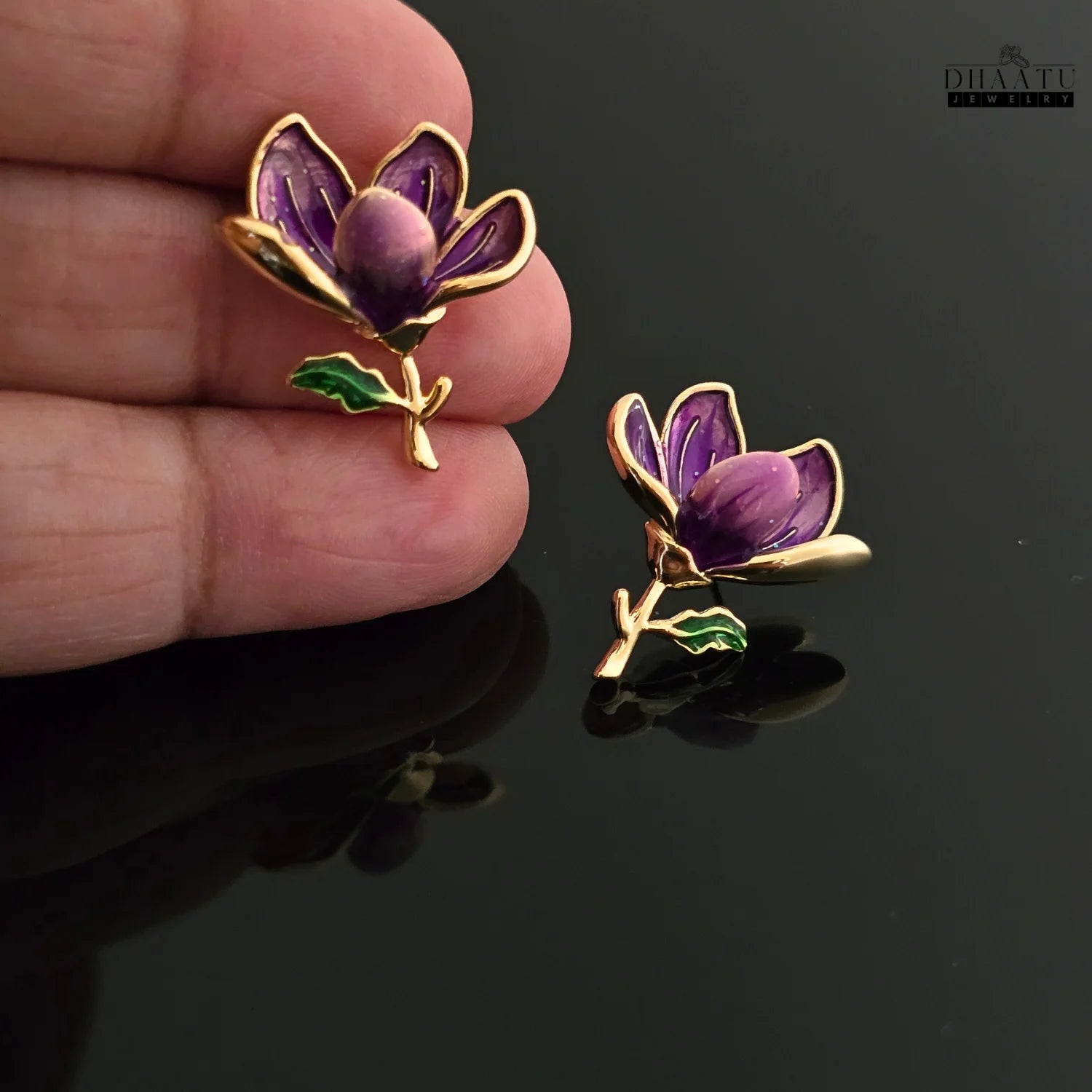 A hand holding one of the Dhaatu Jewelry purple floral stud earrings, with the second earring resting on a black surface, providing a sense of scale and detail.