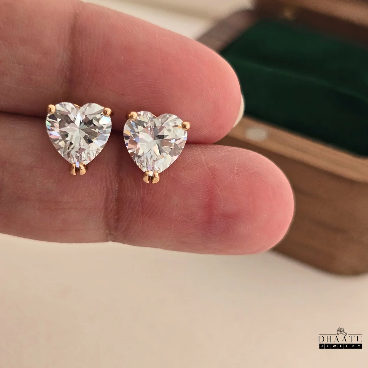 A pair of Dhaatu Jewelry rose gold tone heart-shaped AD stud earrings gently held between fingers, showing their size and brilliance.