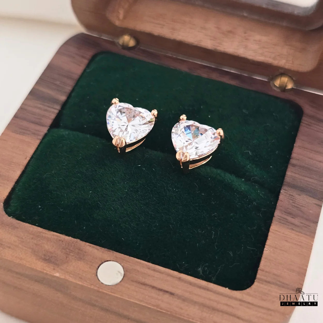 Pair of rose gold finish heart-shaped American Diamond stud earrings from Dhaatu Jewelry, presented in a dark green velvet-lined wooden gift box.