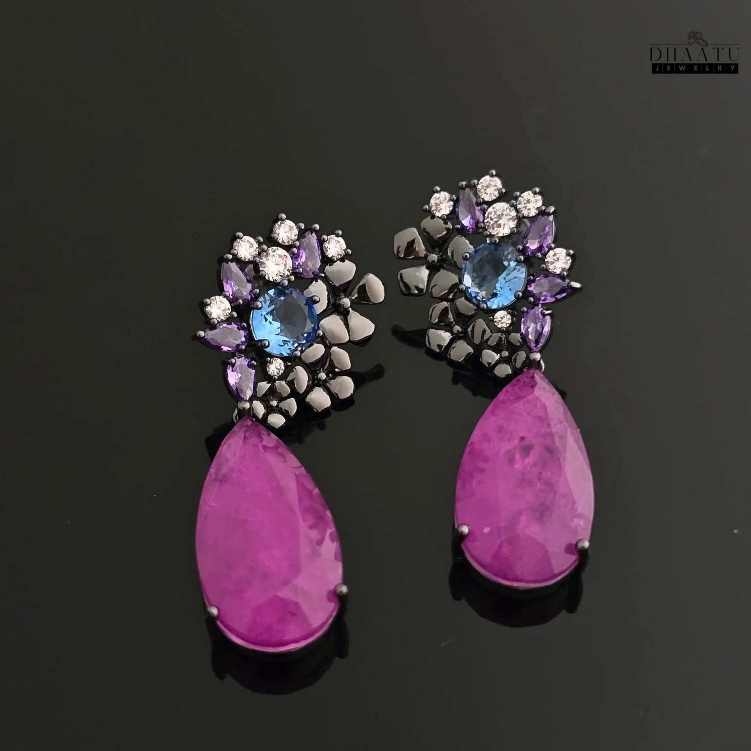 Elegant DHAATU JEWELRY earrings with violet teardrop stones and floral top on a black reflective surface.