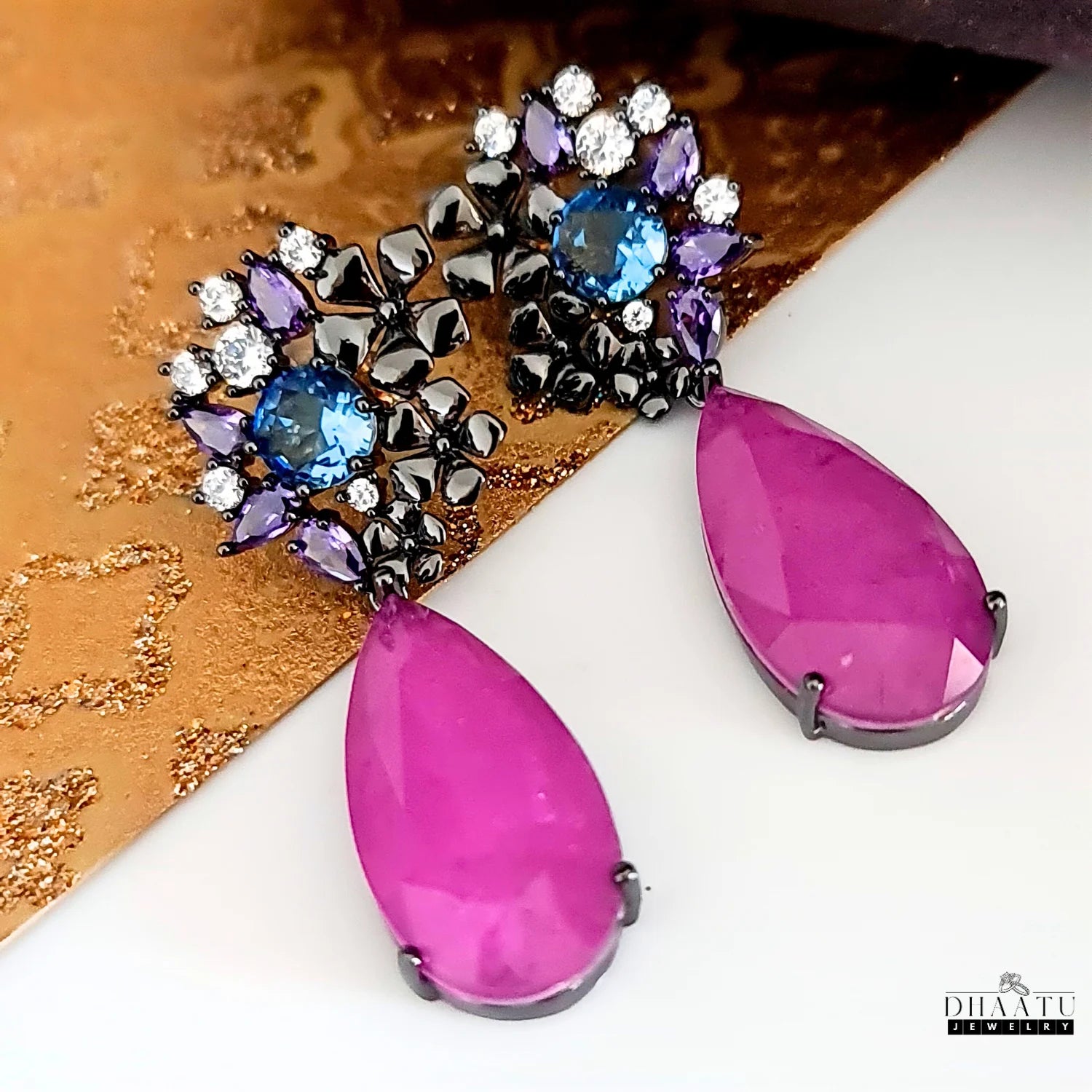 Luxurious DHAATU JEWELRY drop earrings with a floral motif and large violet stones, displayed on a gold and white background.