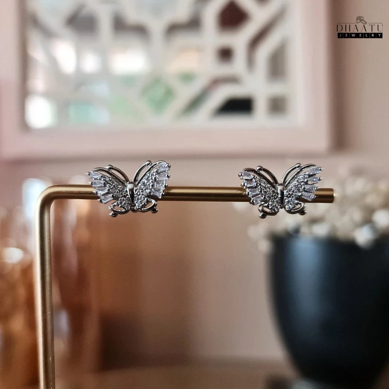Close-up display of Dhaatu Jewelry silver butterfly stud earrings with zircon stones and white gold plating, on a gold stand.