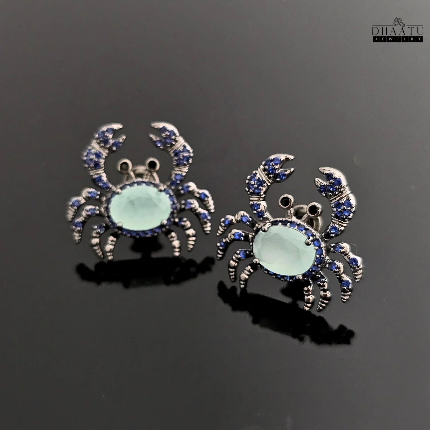 Pair of silver-toned crab earrings with dark blue and aquamarine zircon stones on a glossy black background.