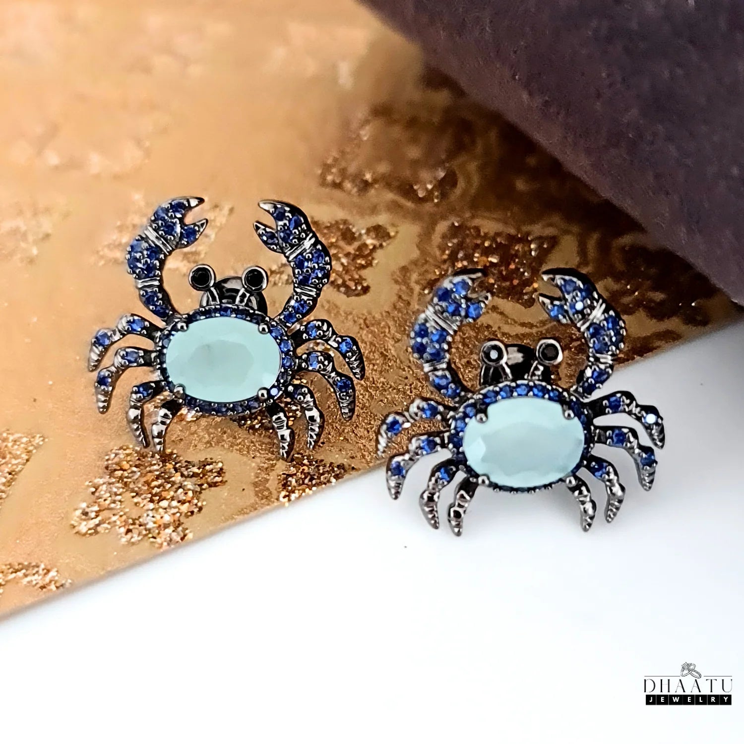 Pair of silver-toned crab earrings with dark blue and aquamarine zircon stones on a textured gold and white background